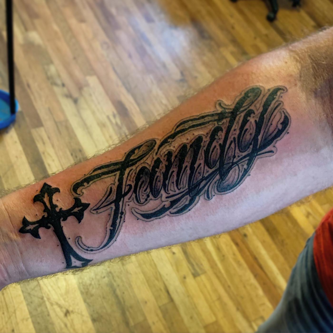 The word Family in ornate script lettering with a gothic cross, on a forearm